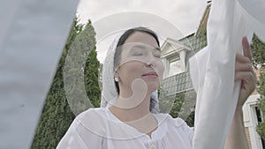 Portrait of attractive senior woman with a white shawl on her head removing bed linen from the rope outdoors close-up