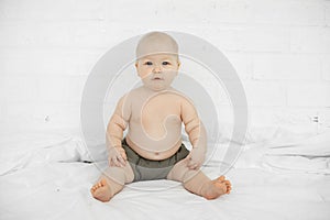 Portrait of attractive serious grey-eyed plump cherubic baby infant toddler wearing grey pants sitting on white bed.