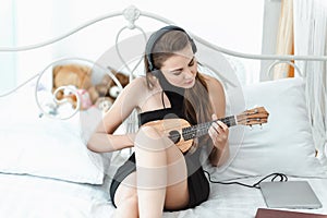 Portrait of Attractive Sexy Woman is Relaxing on Bedroom While Playing Ukulele Instrument and Listen Music on Headphones on