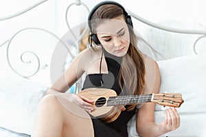 Portrait of Attractive Sexy Woman is Relaxing on Bedroom While Playing Ukulele Instrument and Listen Music on Headphones on