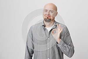 Portrait of attractive smiling bald man with beard showing okay sign and standing with not bad expression over gray