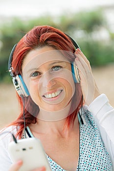 Portrait of an attractive smiling redhead woman listening to music on her phone with greenery in the background