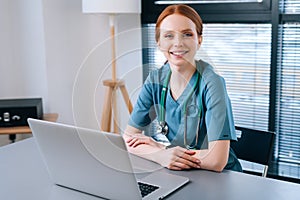 Portrait of attractive smiling young female doctor in blue green medical uniform sitting at desk with laptop on