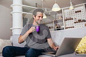 Smiling young man wearing casual clothes sitting on a couch in the living room, using laptop computer