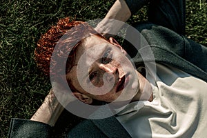 Portrait of attractive stylish young guy model with red hair and freckles sitting on green grass, wearing jacket. Fashionable outd