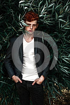 Portrait of attractive stylish young guy model with red hair and freckles sitting on green grass, wearing jacket. Fashionable outd