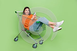 Portrait of attractive trendy cheerful girl riding cart using device smm blogging isolated over bright green color