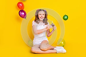 Portrait of attractive trendy cheerful girl sitting on floor with air balls eating small festal cake isolated over