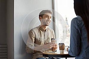 Portrait of attractive unshaved young guy with dark hair, smiling, drinking coffee and listening to girlfriend stories