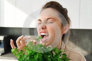 Portrait of attractive woman biting parsley, eating fresh vegetables and herbs in the kitchen, enjoying healthy