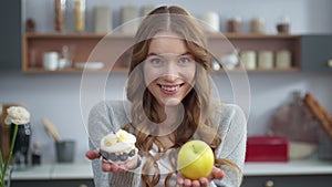 Portrait of attractive woman choosing between cake and apple at home kitchen.