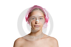 Portrait of attractive woman in pink cap with plasters on nose after rhinoplasty against white studio background.