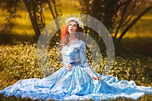 Portrait of an attractive woman with a rim of white flowers and in a blue magnificent dress sitting on the grass in the park again