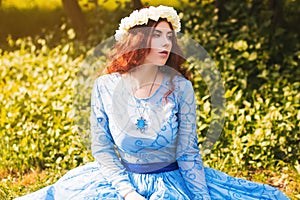 Portrait of an attractive woman with a rim of white flowers and in a blue magnificent dress sitting on the grass in the park again