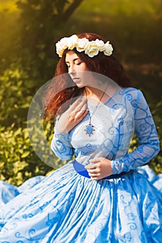 Portrait of an attractive woman with a rim of white flowers and in a blue magnificent dress sitting on the grass in the park again