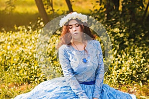 Portrait of an attractive woman with a rim of white flowers and in a blue magnificent dress sitting on the grass in the park again