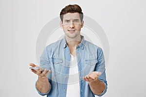 Portrait of attractive young adult with perplexed and confused expression, holding smartphone and showing with gestures