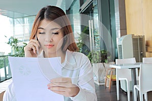 Portrait of attractive young Asian business woman talking on phone against holding document files on her hand in office.