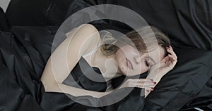 Portrait of attractive young blonde woman sleeping in bed on black pillow and under black blanket. She is talking and