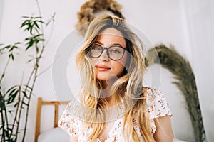 Portrait of attractive young happy caucasian woman in glasses, sitting in cozy bedroom