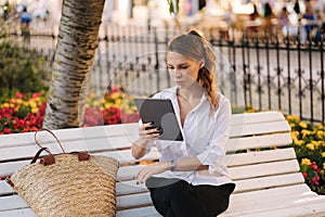 Portrait of attractive young lady in forma outfit sitting on bench in centre of city and using tablet.