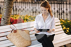 Portrait of attractive young lady in forma outfit sitting on bench in centre of city and using tablet.