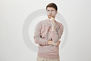 Portrait of attractive young man with bristle looking up while touching chin and thinking about something, standing over