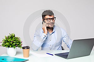 Portrait of attractive young man in glasses sitting at office desk with laptop computer and talking on mobile phone. Communication