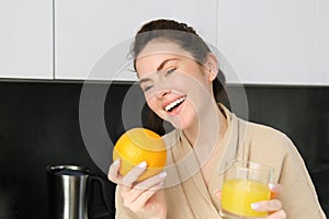 Portrait of attractive young modern woman, posing in kitchen, holding glass of juice and an orange, laughing and talking