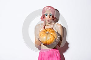 Portrait of attractive young party girl in pink wig looking amazed at pumpkin, celebrating halloween, standing over