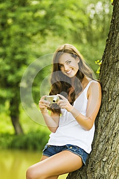 Portrait of attractive young smiling brunette girl wearing white shirt and making photos at summer green park with compact camera.