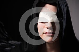 Portrait of an attractive young woman in a hood and black cape with a blindfold. Low key sexy caucasian girl justice and