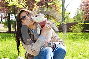 Portrait of attractive young woman hugging cute jack russell terrier puppy in park, green lawn, foliage background. Hipster female