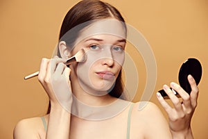 Portrait of attractive young woman with long brunette hair looks at camera and applies cosmetic product against beige