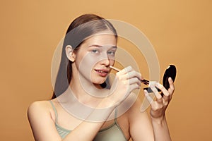 Portrait of attractive young woman with long brunette hair looks at camera and holds cosmetic product against beige