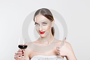 Portrait of attractive young woman, looking at camera, holding a glass of red wine and showing thumbs up, isolated on white wall