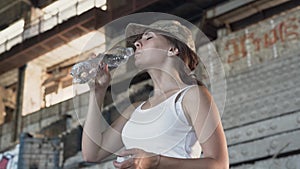 Portrait of attractive young woman in military cap drinking water from the bottle in dusty dirty abandoned building