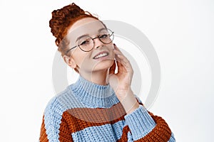 Portrait of attractive young woman with red hair combed in hairbun, touching her natural pale facial skin and smiling