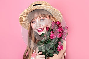 Portrait of an attractive young woman in summer dress and straw hat holding bouquet of roses on pink background