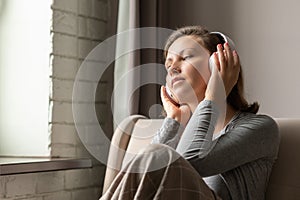 Portrait of Ñaucasian young woman in headphones listening music sitting on sofa covered blanket, drinking coffee from cup