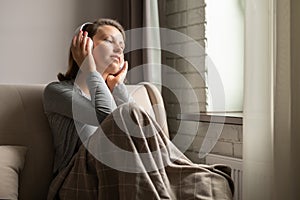 Portrait of Ñaucasian young woman in headphones listening music sitting on sofa covered blanket, drinking coffee from cup