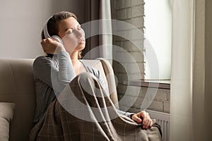 Portrait of Ñaucasian young woman in headphones listening music sitting on sofa covered blanket, drinking coffee from cup