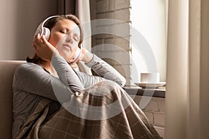 Portrait of Ñaucasian young woman in headphones listening music sitting on sofa covered blanket, drinking coffee from cup