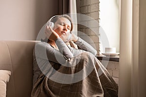 Portrait of Ñaucasian young woman in headphones listening music sitting on sofa covered blanket, drinking coffee from cup