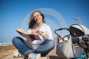 Portrait of authentic mother reading book outdoor, spending nice time on the beach with her baby sleeping in stroller