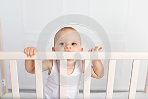 Portrait of a baby boy 8 months old standing in a crib in a children`s room in white clothes and nibbling on the bed, baby`s