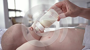 Portrait of baby girl drinking milk from a bottle that mother holds in her hands, newborn in diaper is lying on changing