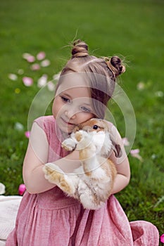 portrait baby girl in a pink dress plays with a rabbit in a green meadow in summer. Funny friendship between a child and