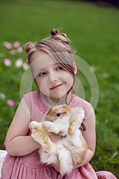 portrait baby girl in a pink dress plays with a rabbit in a green meadow in summer. Funny friendship between a child and
