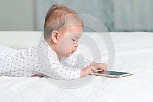 Portrait of baby is lying on the bed and playing with a smartphone. Side view. Copy space. Concept of children`s games with moder